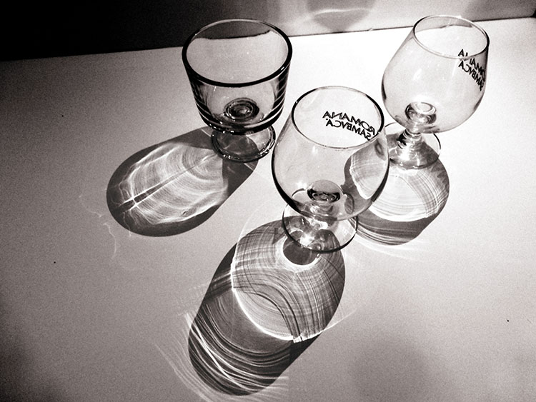 Three glasses standing in front of a light. The camera views them from above, capturing their shining shading cast across the white page below, upon which those glasses stand.
